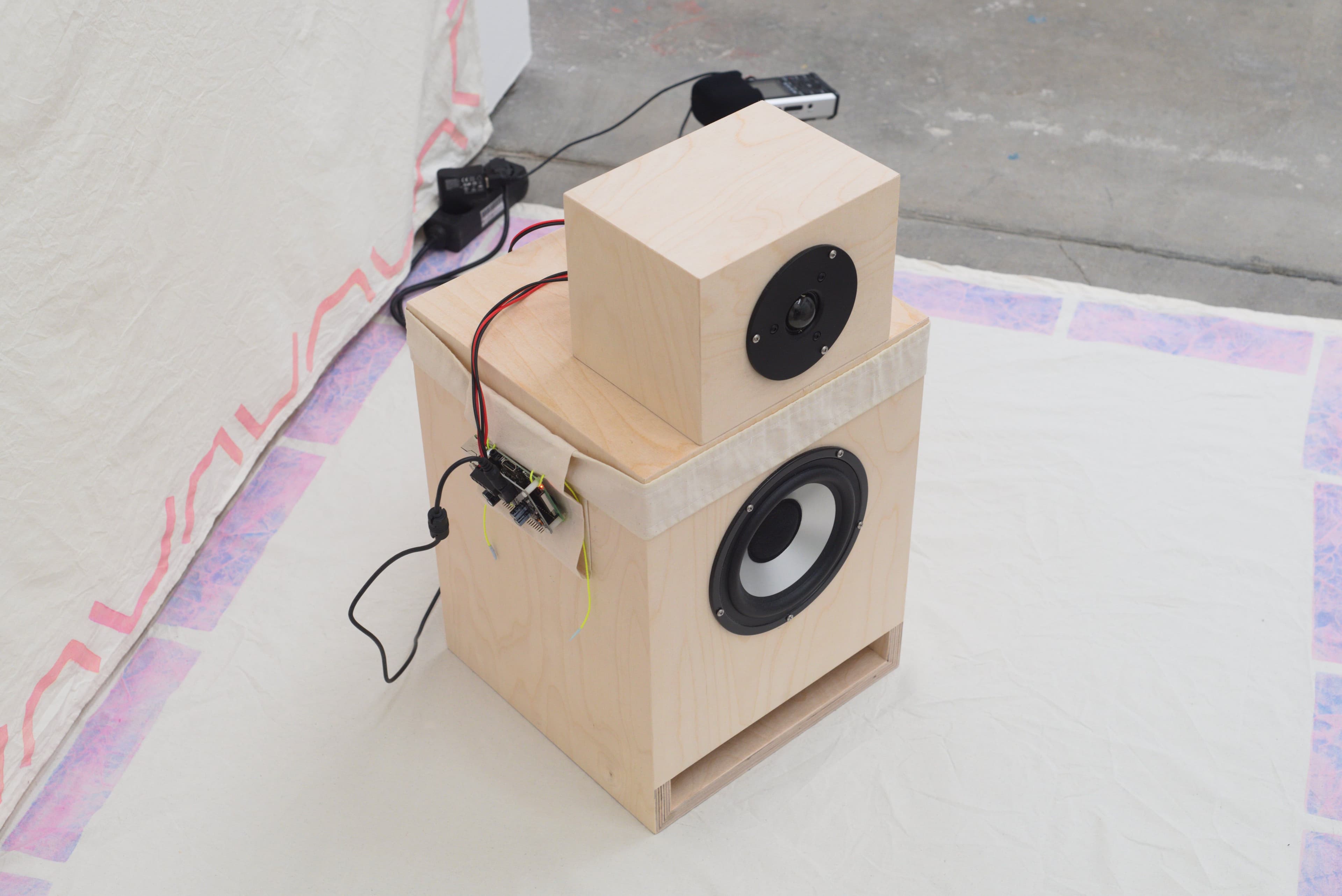 A speaker plugged in, in a gallery setting, with hand painted cotton works behind and under it