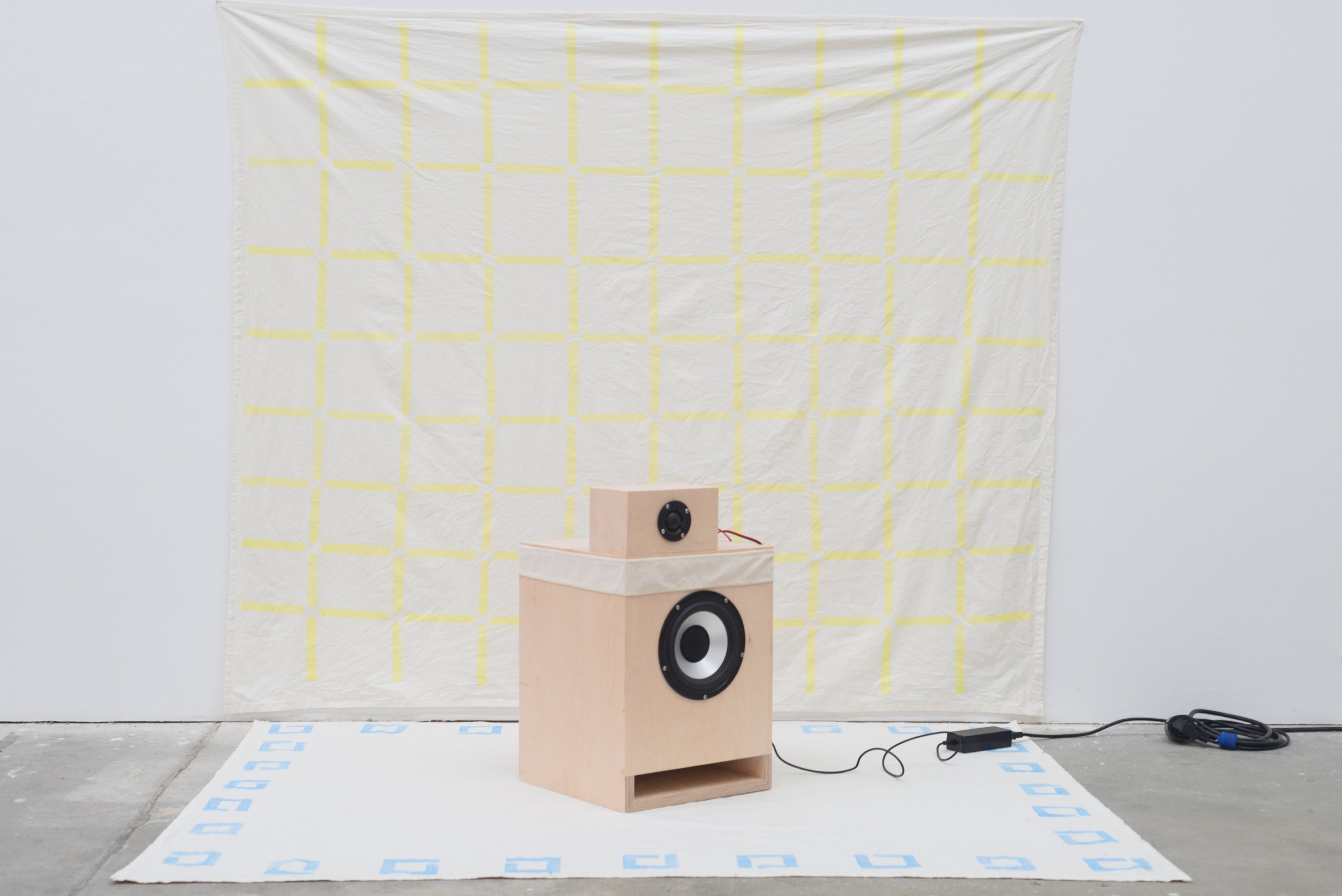 A speaker plugged in, in a gallery setting, with hand painted cotton works behind and under it